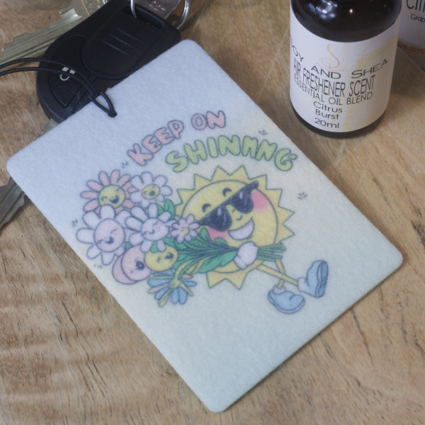 A rectangular car air freshener is rests on a car key next to a bottle of air freshener scent.  The Air Freshener has a cartoon image of a smiling sun walking along with a happy bunch for flowers with the text KEEP ON SHINING against an ombre background of blue to green.