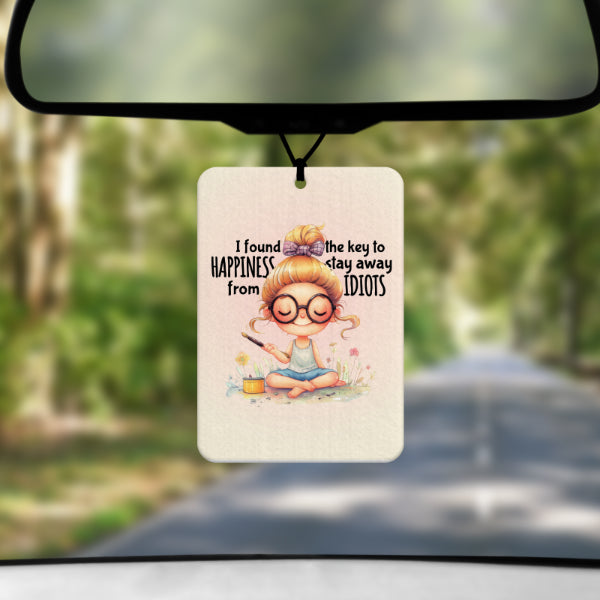 A mock up image showing a view through a car windscreen with a long tree lined road.  From the rearview mirror hangs an air freshener with a drawing of a young woman sitting cross legged on some grass with some flowes and incense.  Around her head are the words  I FOUND THE KEY TO HAPPINESS.  STAY AWAY FROM IDIOTS