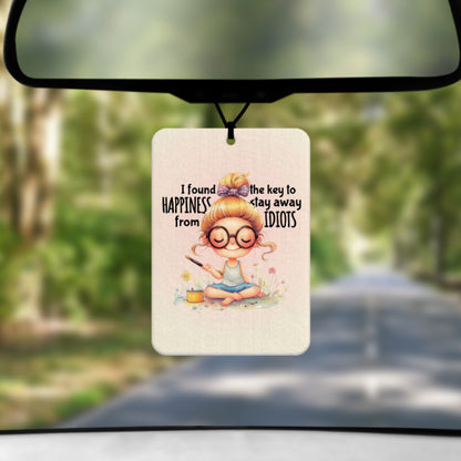 A mock up image showing a view through a car windscreen with a long tree lined road.  From the rearview mirror hangs an air freshener with a drawing of a young woman sitting cross legged on some grass with some flowes and incense.  Around her head are the words  I FOUND THE KEY TO HAPPINESS.  STAY AWAY FROM IDIOTS