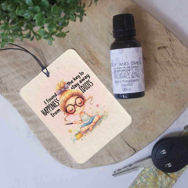 A rectangular car air freshener lies on a wooden surface next to a glass bottle of Air Freshener Scent and car key.  The Air Freshener has a watercolour drawing of a young women sitting cross legged on some grass surrounded by flowers and holding an incense stick.  Around her head are the words I FOUND THE KEY TO HAPPINESS.  STAY AWY FROM IDIOTS