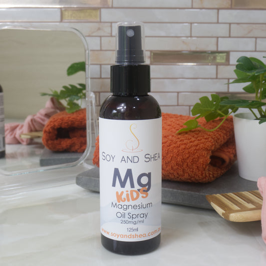 An amber bottle with atomiser lid stands on a marble surface against a tiled background with a facecloth, plant and mirror in background.  The bottle has a white label idenitifying it has Magnesium Oil Spray with the word KIDS in a bright orange, fun font in bold.