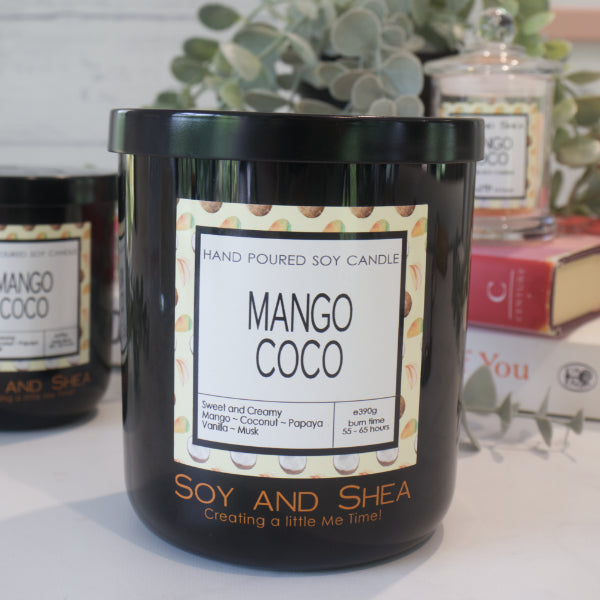 A black candle jar with a lid embossed with the Soy and Shea Logomark sits on a white counter with a label reading MANGO COCO and 390g, sits on white counter with a stack of candles in the background