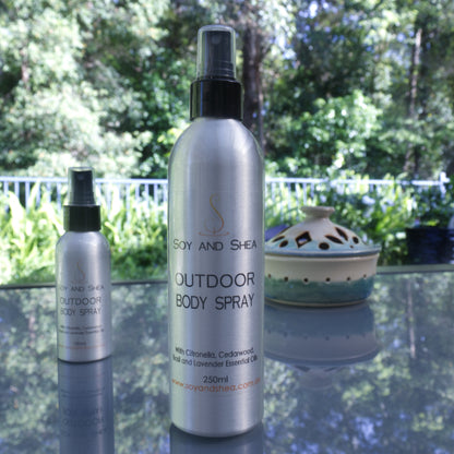 Two metal bottles of "Outdoor Body Spray" by Soy and Shea, stand on a glass table top with a lush green garden in the background.  A large bottle sits in the front of the images with an atomiser style lid and reads 250ml, while a smaller bottle sits towards the back slightly blurred.