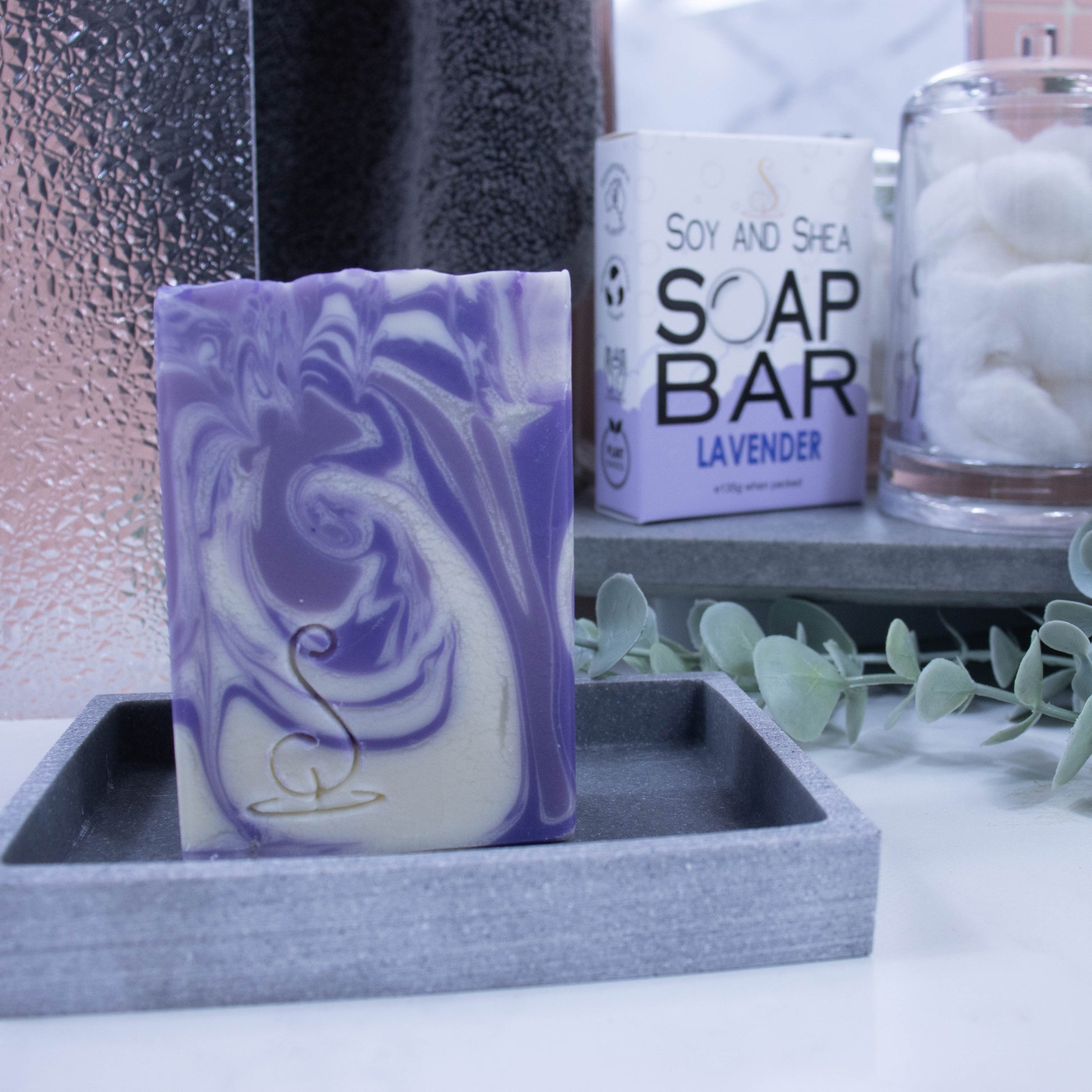 A tall rectangular bar of soap sits on a grey soap dish. The soap features swirls of colour in white, lavender and dark purple.  Behind the soap is a shower screen and towel along with a box the soap was presented in.  The box has a white top with pale purple bubble border.