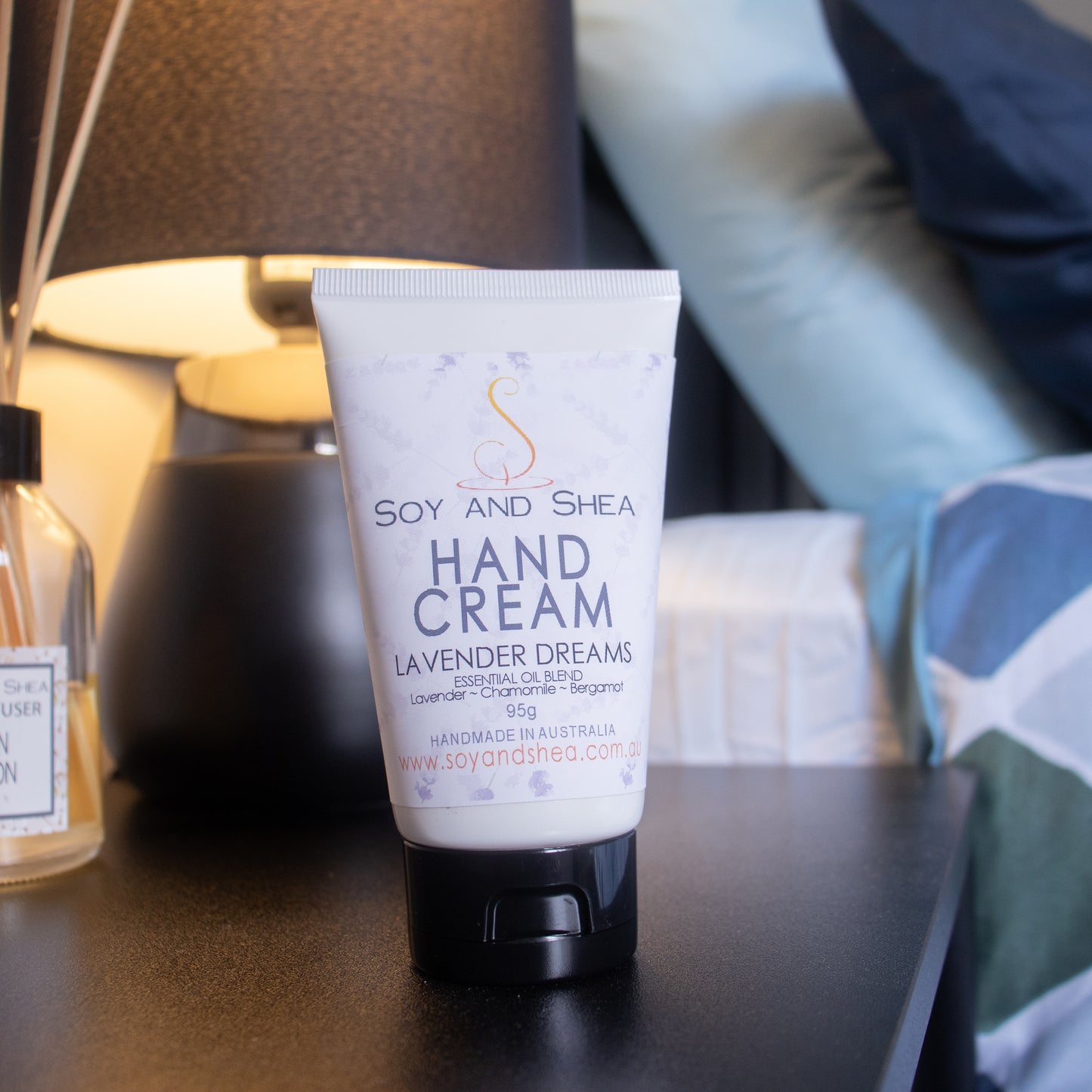 A tube of Soy and Shea Hand Cream labeled "Lavender Dreams" is placed on a bedside table. The table also holds a bottle and a lamp, with the blurred background showing pillows and part of a bed. The hand cream tube is prominently displayed in the foreground and is white with a black lid with a white label with purple lavender sprigs