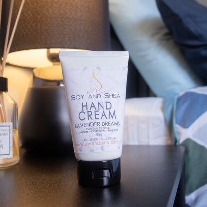 A tube of Soy and Shea Hand Cream labeled "Lavender Dreams" is placed on a bedside table. The table also holds a bottle and a lamp, with the blurred background showing pillows and part of a bed. The hand cream tube is prominently displayed in the foreground and is white with a black lid with a white label with purple lavender sprigs