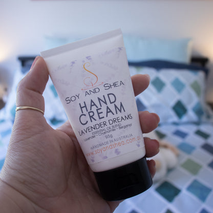 A hand holds a white tube with black lid and a white label reading HAND CREAM LAVENDER DREAMS against a blurred background of a bed