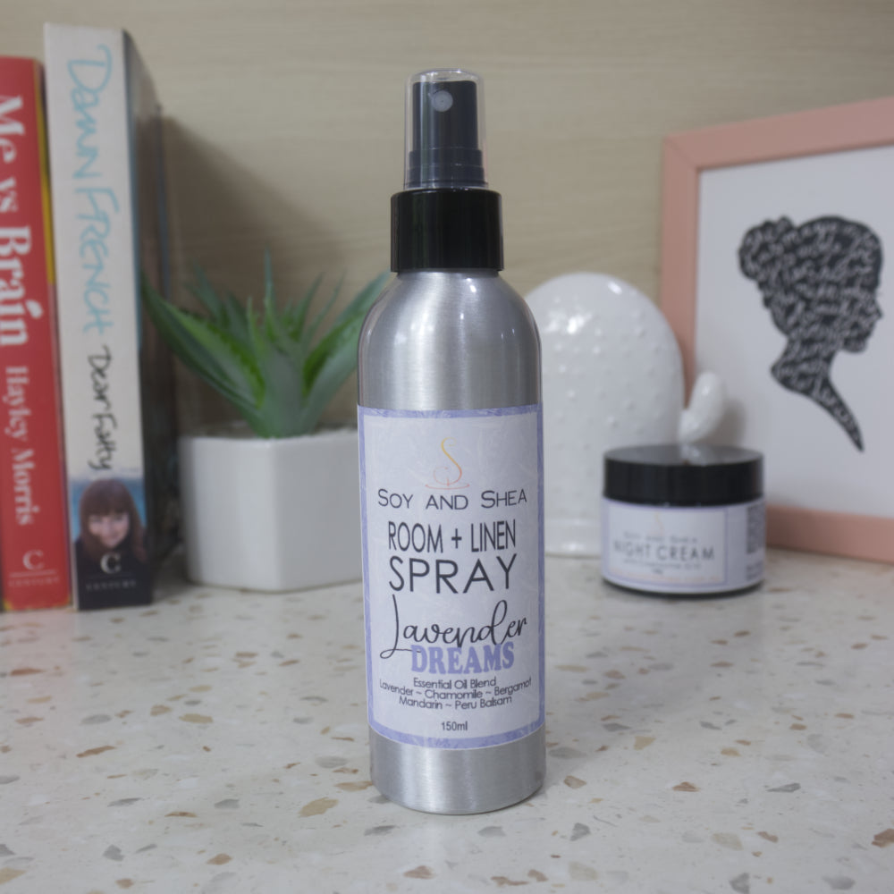 A silver metal bottle with black atomiser sprayer stands on a terrazzo surface against a background with a books, plant and picture frame.   The bottles features a white label with purple frame with product details.
