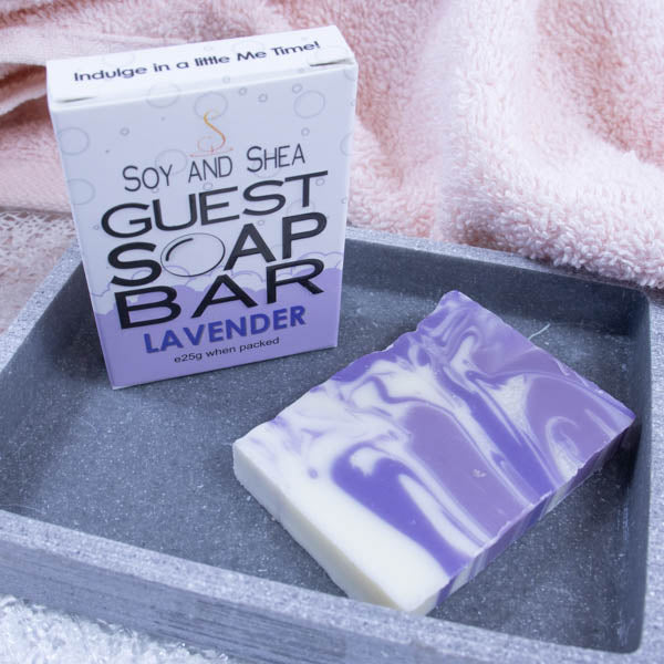 A grey stone soap dish sits next to a pink towel.  On the dish stands a white box with a border of purple bubble foam across the bottom and text GUEST SOAP BAR. Next to it lies a small piece of soap that has a white base with swirls of purple throughout