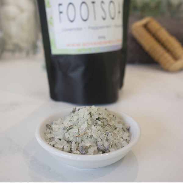 A small white dish sits on a marble counter with a blurred background showing the pouch of Foot Soak.  The bowl is filled with white salts speckled with purple lavender heads and dried green leaves. 