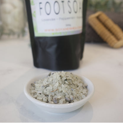 A small white dish sits on a marble counter with a blurred background showing the pouch of Foot Soak.  The bowl is filled with white salts speckled with purple lavender heads and dried green leaves. 