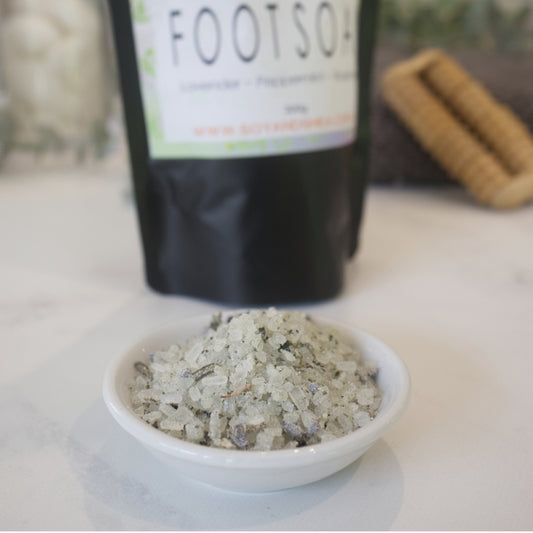 A small white dish sits on a marble counter with a blurred background showing the pouch of Foot Soak.  The bowl is filled with white salts speckled with purple lavender heads and dried green leaves. 