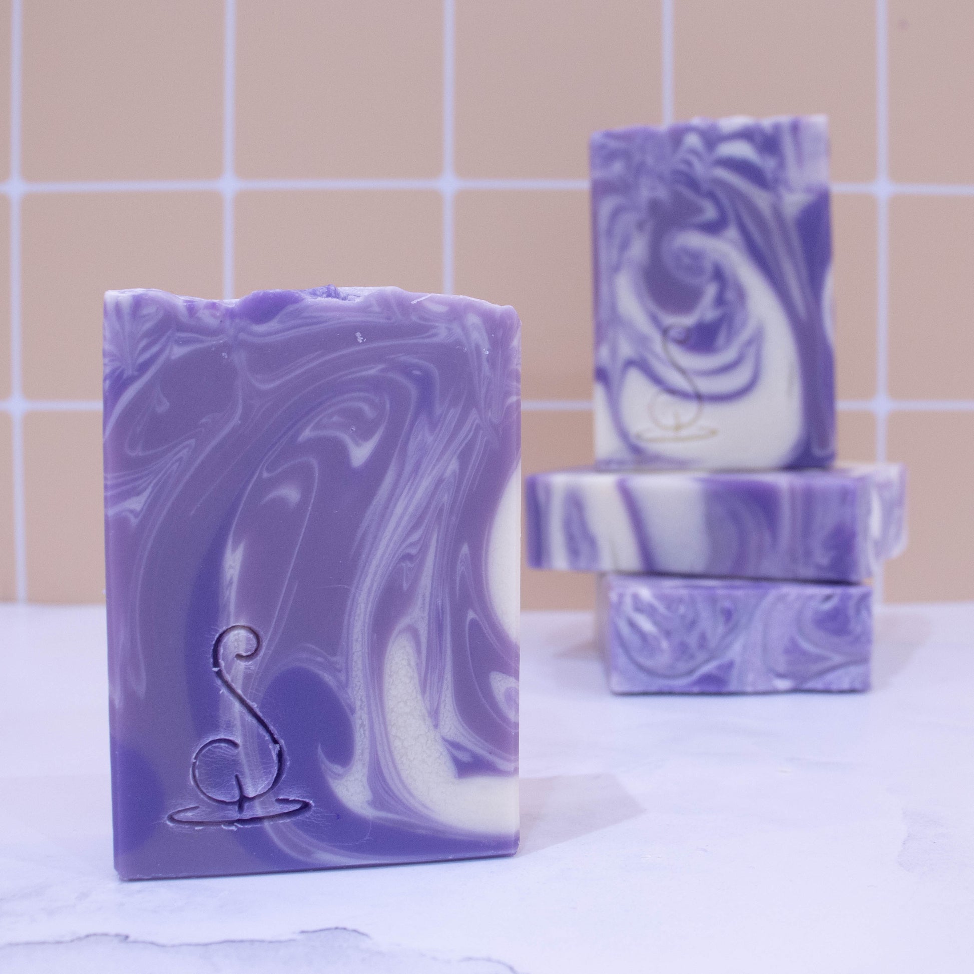 Four rectangular bars of soap sit on a marble bench each showing various design patterns from the same batch of soap. Two soaps lay flat but stacked upon each other showing a flat top.  A third soap stands on these while a fourth is presented in front of the stack.   All soaps feature a swirling pattern of white, lavender and dark purple soap but all have slight differences