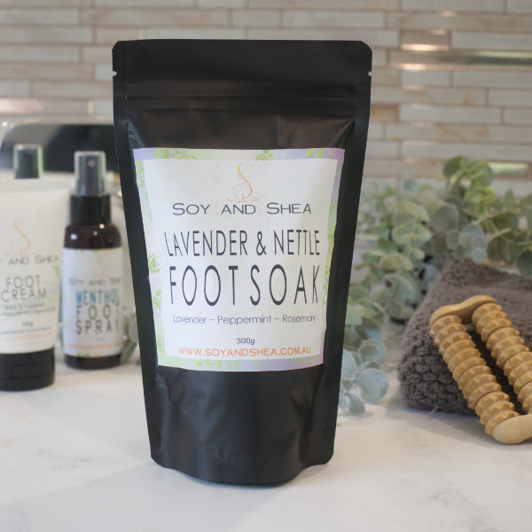 A black zip lock pouch of Soy and Shea Lavender and Nettle Foot Soak sits on a bathroom counter with Foot Cream and a bottle of Foot Spray in the background against a decorative tile wall and greenery
