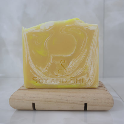 Yellow soap bar with white swirls and 'Soy and Shea' branding on a wooden stand against a white background