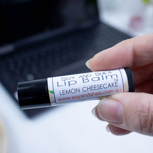 A hand holds a black tube of lip balm against a blurred background with an open laptop computer on a white surface.  The label has text LIP BALM LEMON CHEESECAKE on a white label with a border showing illustrations of cheesecake slices.