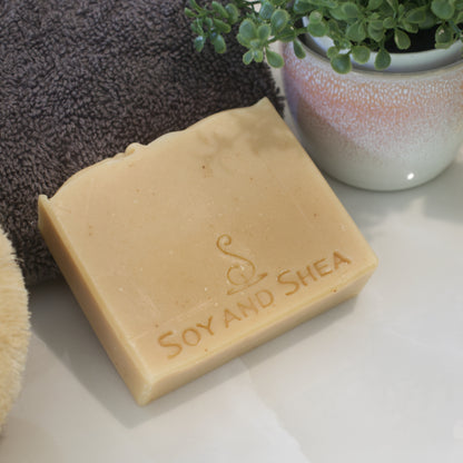 A pale yellow soap with dark yellow flecks rests agains a dark grey towel beside a potted plant on white surface.