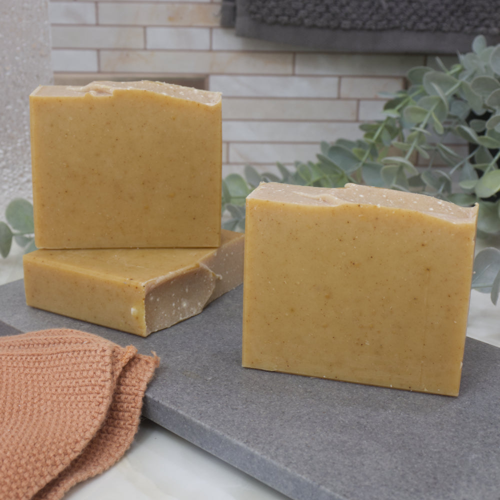 Three bars of yellow soap on a gray surface with a textured towel and greenery in the background.