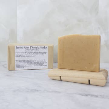 A pale yellow soap bar with dark yellow flecks sits on a wooden stand with a brown paper package behind labeled 'Lemon, Honey and Turmeric Soap Bar' on a marble background
