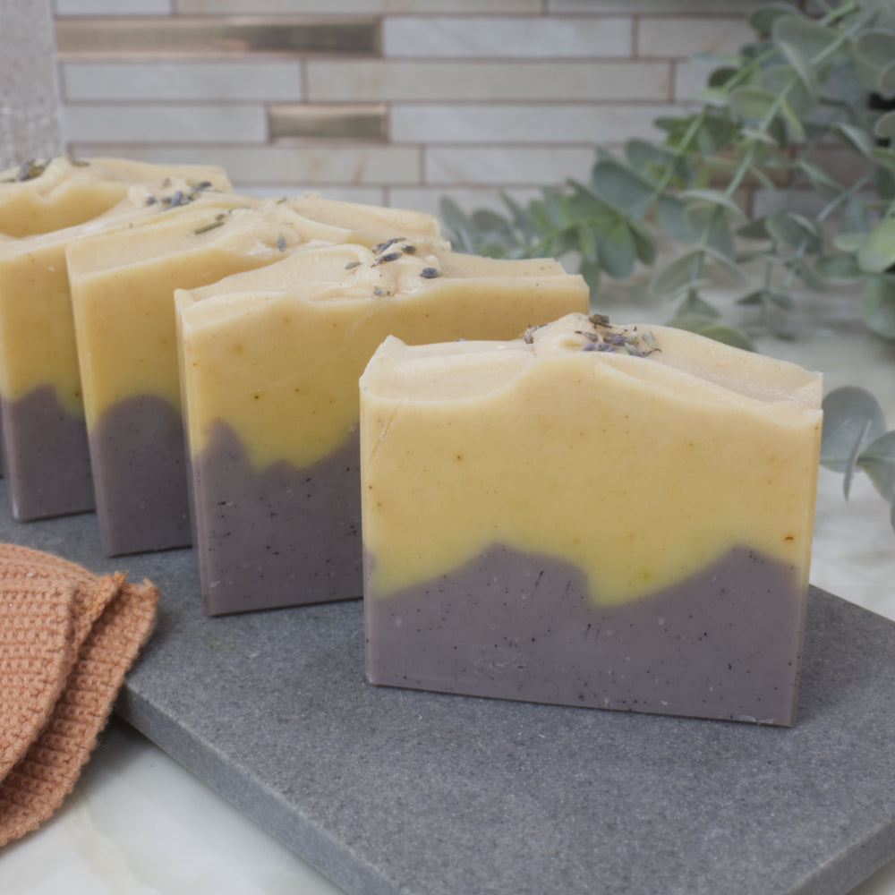 Four soap bars in a row on a gray stone surface, with a plant in the background.  The soaps feature a layered pattern with dark purple base and bright yellow top.   Lavender pieces can be seen on top of the soaps