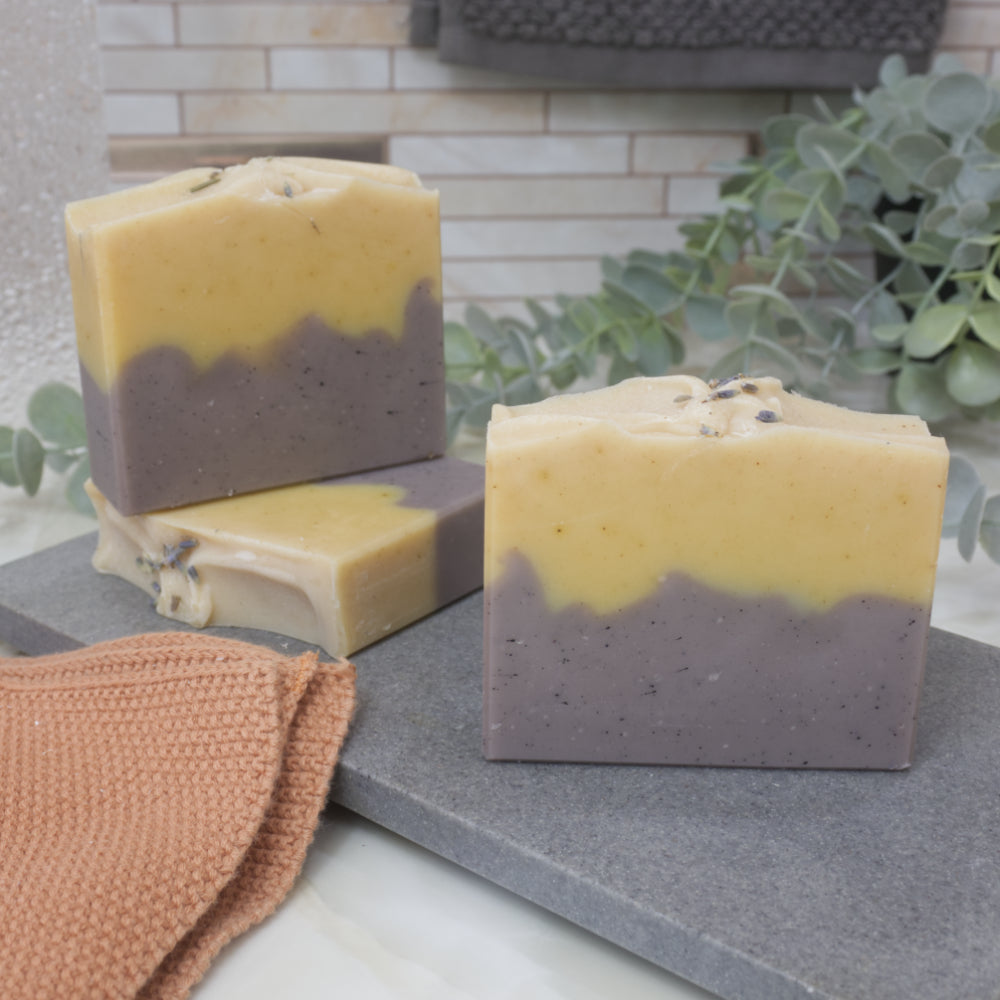 Three soap bars sit on a gray stone surface with a plant in the background. The soaps feature a layered pattern with dark purple base and bright yellow top.   Lavender pieces can be seen on top of the soaps