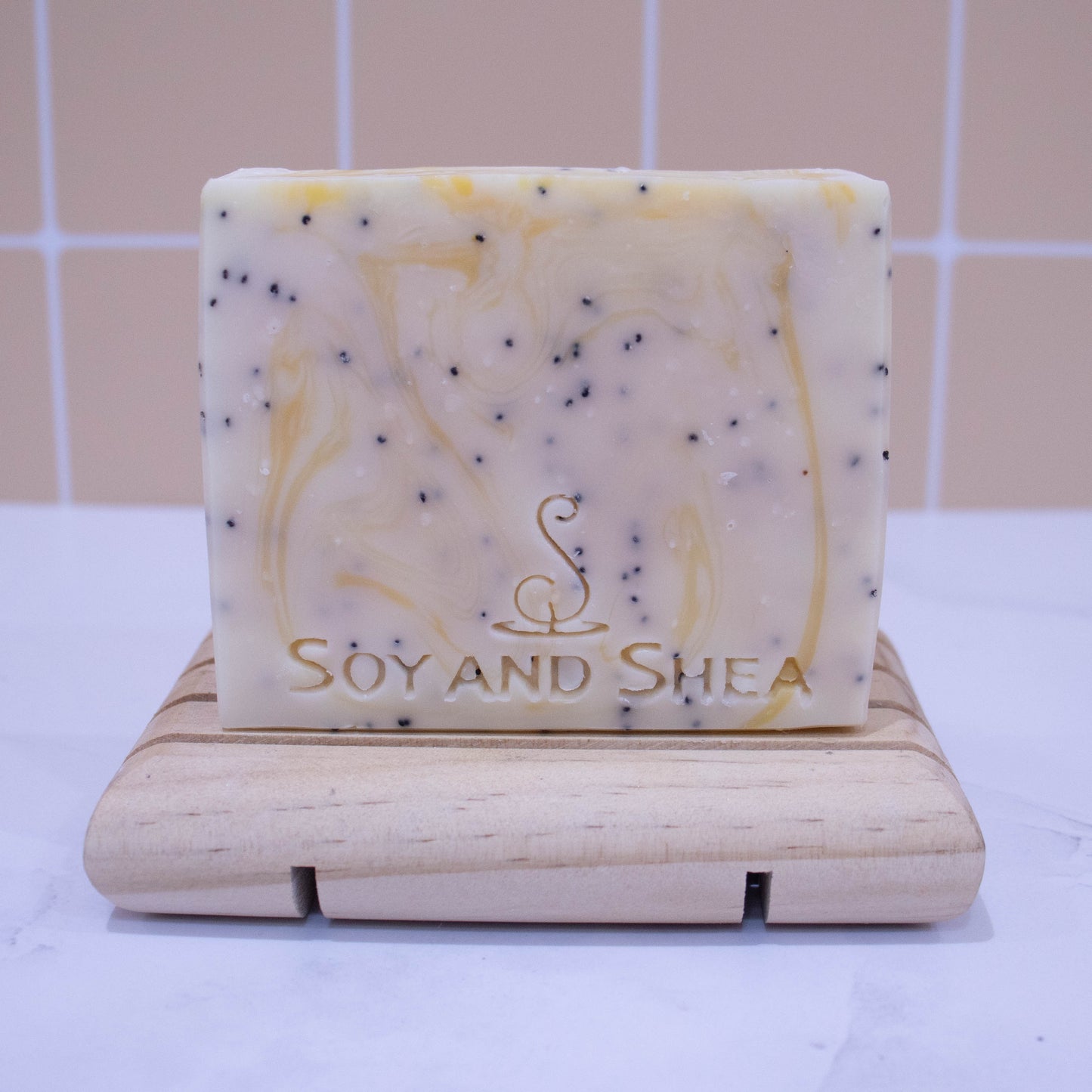 A bar of soap stands on a wooden soap dish on a white surface with peach tile background.  The soap is white with yellow wisps of colour and black flecks of poppy seeds.