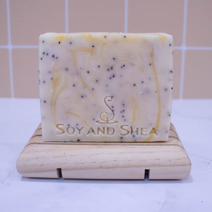 A bar of soap stands on a wooden soap dish on a white surface with peach tile background.  The soap is white with yellow wisps of colour and black flecks of poppy seeds.