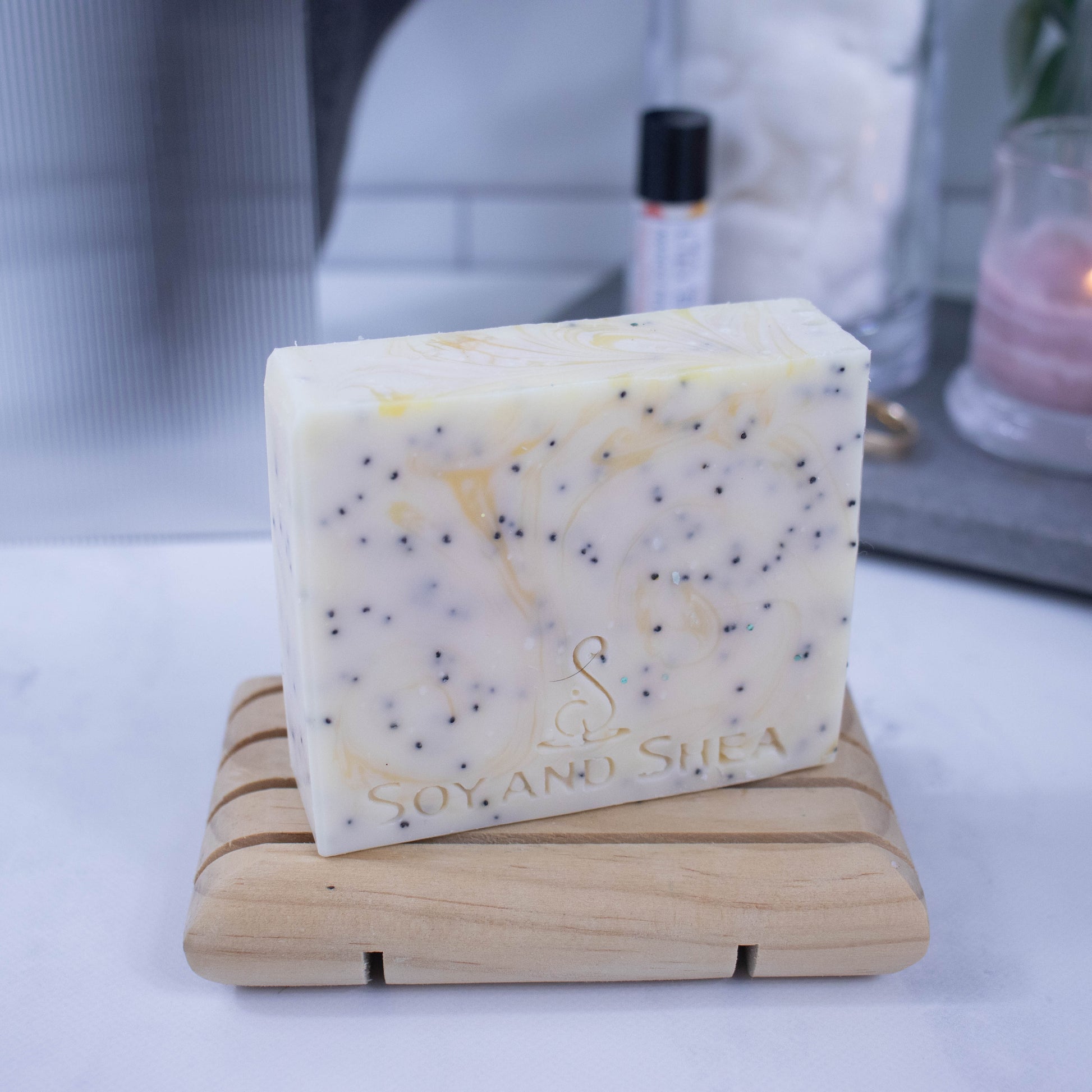 A bar of soap stands on a wooden soap dish displayed on a bathroom countertop with a blurred background.  The soap is yellow with wisps of yellow and flecks of black from poppy seeds.