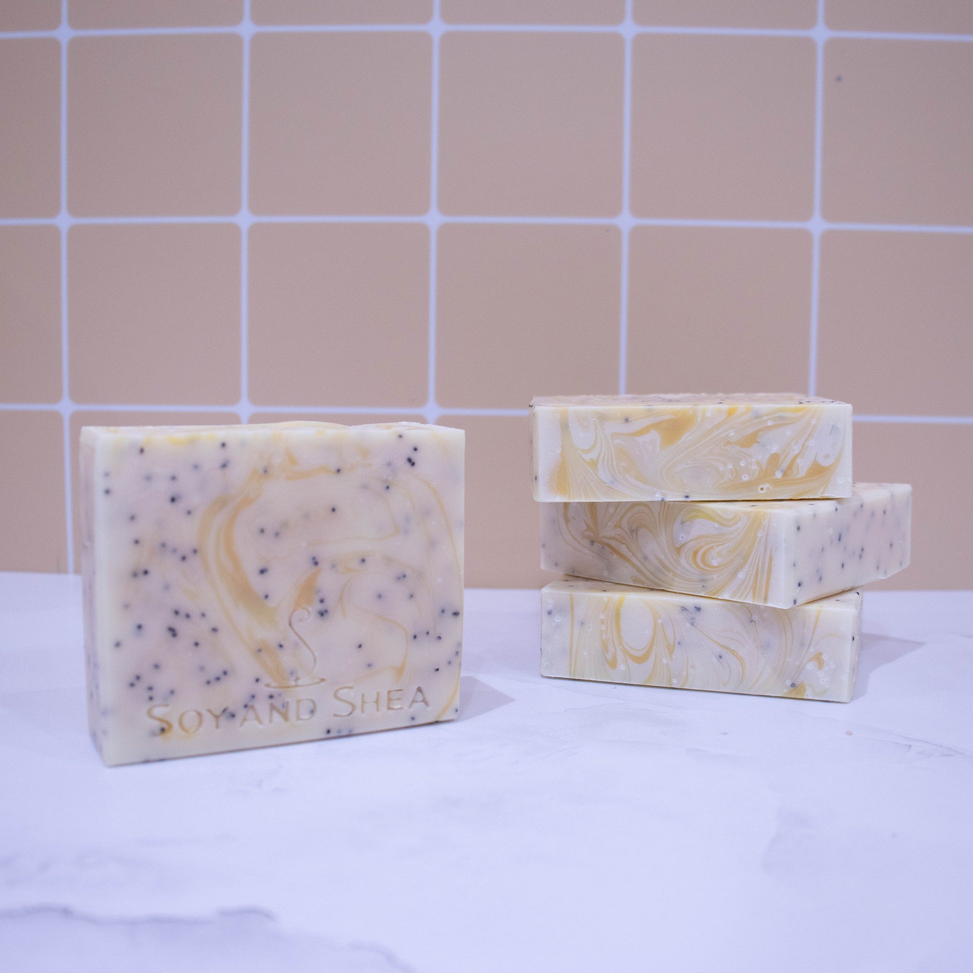 Three soap bars lay stacked upon each other showing a flat while top with yellow swirl.  To the left stands a fourth bar which is white with yellow wisps of colour and black flecks of poppy seeds.  These sit on a marble surface with pale orange tile background.