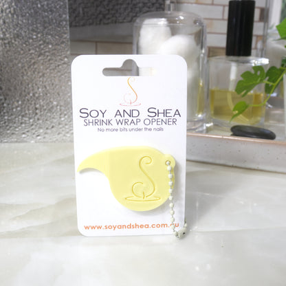 A pale yellow Shrinkwrap opener with matching ball and chain keyring is attached to a white card with product details.  The card is standing upright on a bathroom countertop with a bottle of perfume, a jar of cotton balls and towel in the background