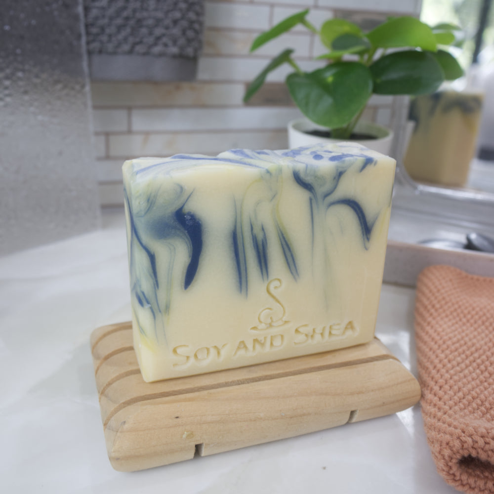 A bar of handmade soap stands on a wooden soap dish on a bathroom countertop.  The soap has a base colour of pale yellow and features blue and yellow streaks of colour.