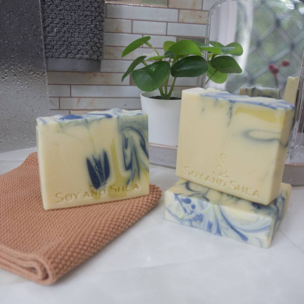 Three bars of soap are presented on a bathroom counter with a face cloth and plant.  One soap stands upright to the left on an orange cloth  while another lays flat to the front right with a third standing on it.  The soaps have a light yellow base colour with drops of blue and dark yellow colouring