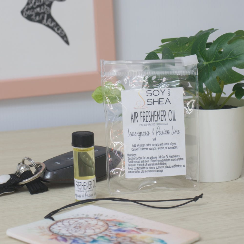 A small clear bottle with black cap of Soy's Shea Air Freshener Oil next to a clear resealable pouch, displayed on a light wood tabletop with a car key, a felt air freshener, and a potted plant in the background.  