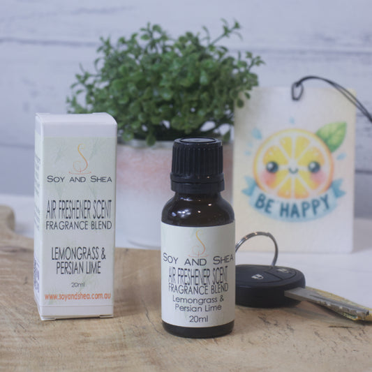 A small amber bottle with black lid labelled AIR FRESHENER SCENT stands on a wooden surface next to the box packaging.  In the background is a car key, air freshener and greenery.