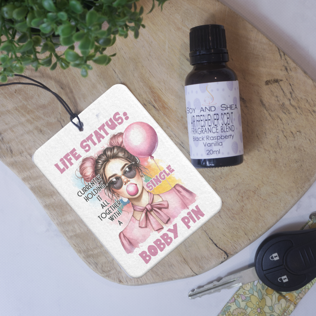 A white car air freshener with a design featuring a cartoon girl holding a pink ballon and blowing pink bubbleum, and text that reads 'LIFE STATUS: CURRENTLY HOLDING IT ALL TOGETHER WITH A SINGLE BOBBY PIN'. The air freshener is displayed in a location that includes a plant and various items on a surface.