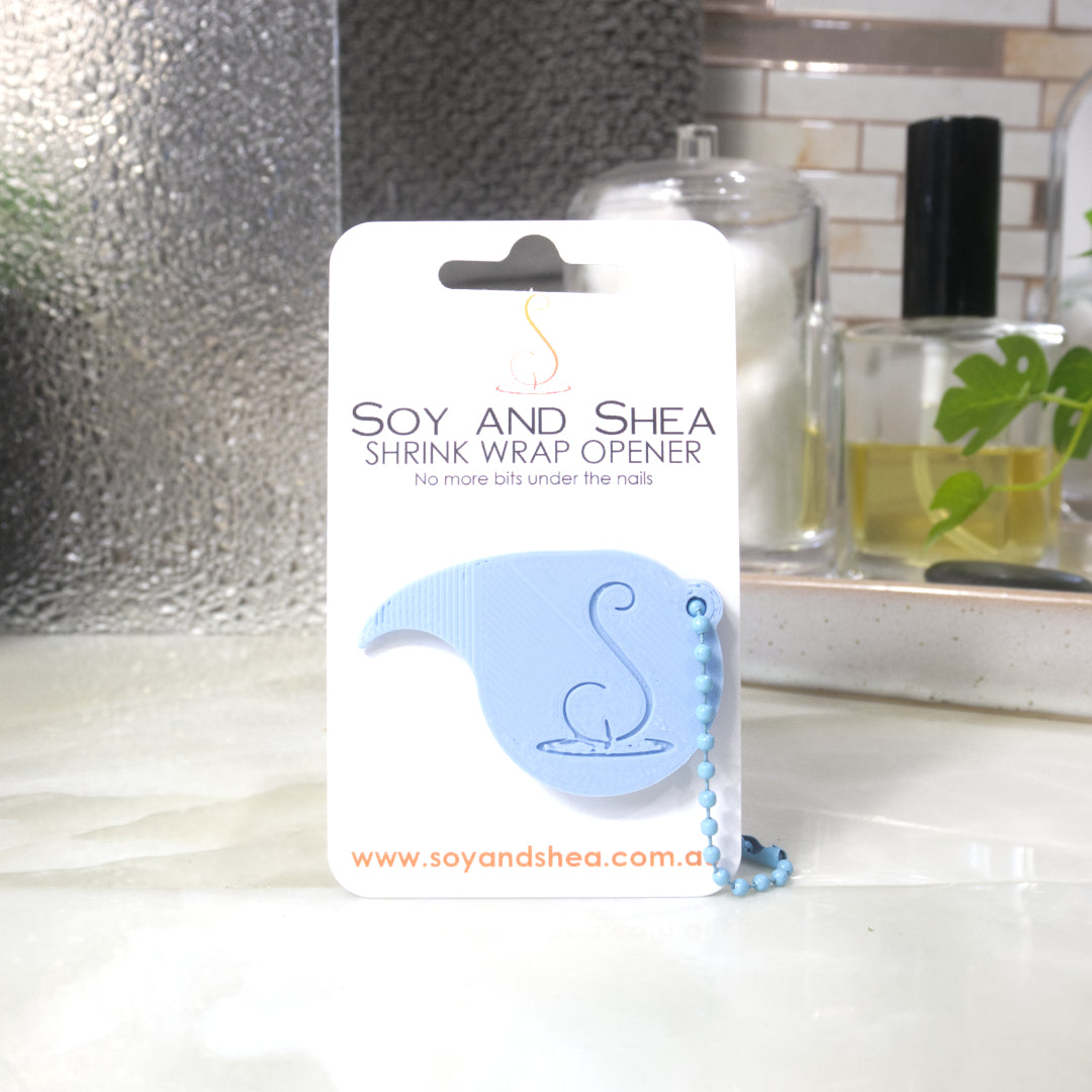 A Light Blue Shrinkwrap opener with matching ball and chain keyring is attached to a white card with product details.  The card is standing upright on a bathroom countertop with a bottle of perfume, a jar of cotton balls and towel in the background