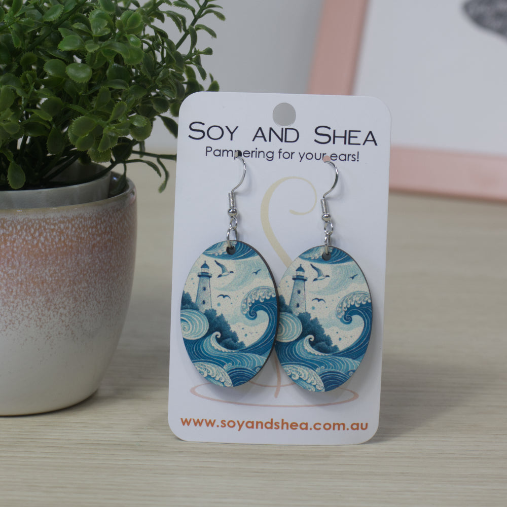 Oval shapped Soy and Shea earrings with wave design featuring a lighthouse  above them on a card, next to a potted plant.
