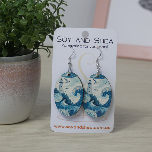 Oval shapped Soy and Shea earrings with wave design featuring a lighthouse  above them on a card, next to a potted plant.