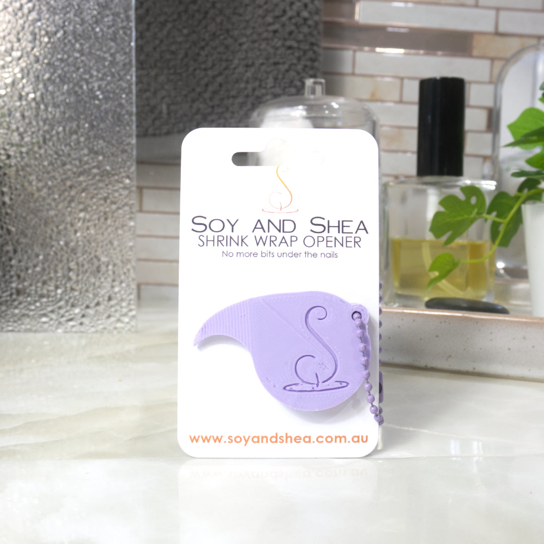 A Lilac Shrinkwrap opener with matching ball and chain keyring is attached to a white card with product details.  The card is standing upright on a bathroom countertop with a bottle of perfume, a jar of cotton balls and towel in the background