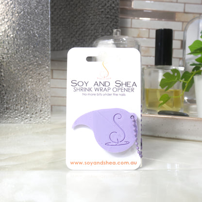 A Lilac Shrinkwrap opener with matching ball and chain keyring is attached to a white card with product details.  The card is standing upright on a bathroom countertop with a bottle of perfume, a jar of cotton balls and towel in the background