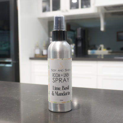 A aluminium bottle with a black atomiser lid stands on a grey kitchen bench agains a blurred background of another kitchen bench with coffee maker.  The bottles features a decorative product label announcing it as Room + Linen Spray and scented in Lime, Basil and Mandarin.