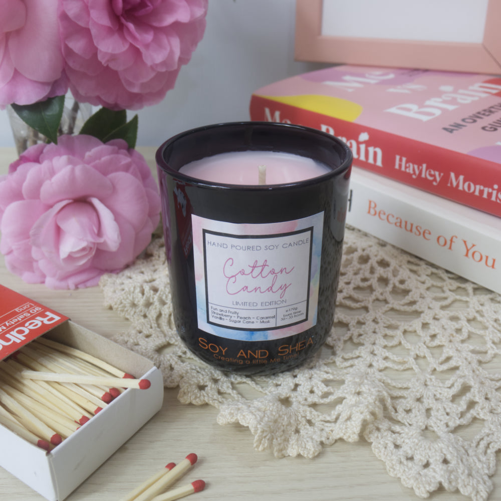 A black candle jar with decorative label is on a sideboard with vase of pink flowers to the left and a stack of bookes to the right.   The lid of the jar has been removed and placed to the right in front of the books revealing a soft pink wax.