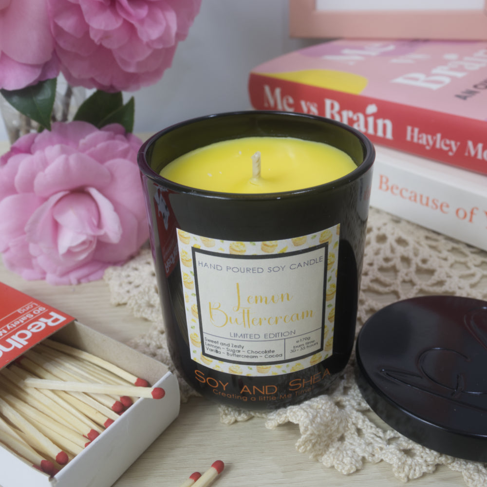 A black candle jar with decorative label is on a sideboard with vase of pink flowers to the left and a stack of bookes to the right.   The lid of the jar has been removed and placed to the right in front of the books revealing a bright yellow wax.