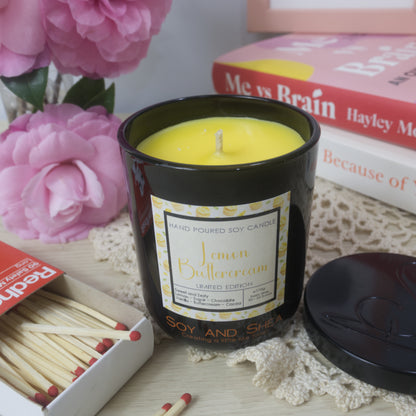 A black candle jar with decorative label is on a sideboard with vase of pink flowers to the left and a stack of bookes to the right.   The lid of the jar has been removed and placed to the right in front of the books revealing a bright yellow wax.