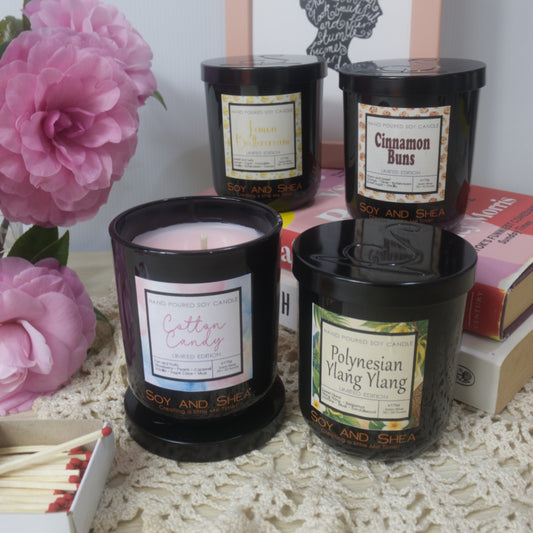 A collection of four candles in black jars are presented on a sideboard next to a vase of pink flowers and box of matches.  Two candles are to the front with one candle having its black lid removed revealing pink wax.  Two more candles are behind these resting on a stack of books.  Each candle is a different scent.