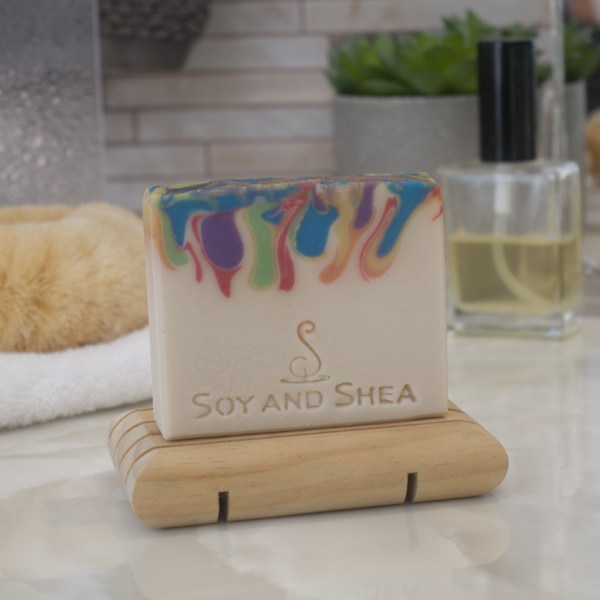 A handcrafted bar of soap with a colorful, swirled design on a wooden soap dish, sits on a bathroom counter top with perfume and body brush in teh background.  The soap has a cream base layer with drips of rainbow colours coming down from the top.