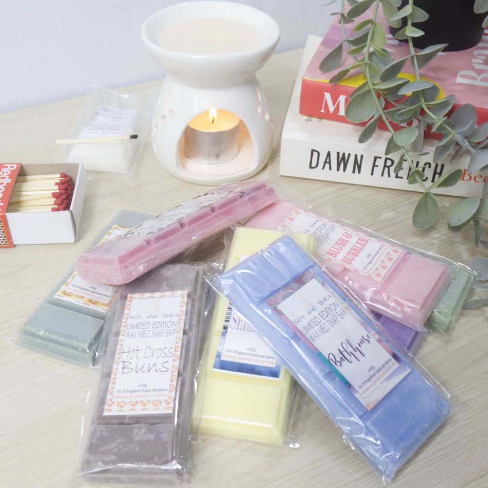 A selection of random wax melts in various colours are piled on a wooden bench next to an oil burner with a lit tea light and a pile of books
