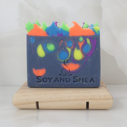 A dark blue soap bar with neon coloured drops and decorative piped top, sits on a wooden soap dish with a marble tile background