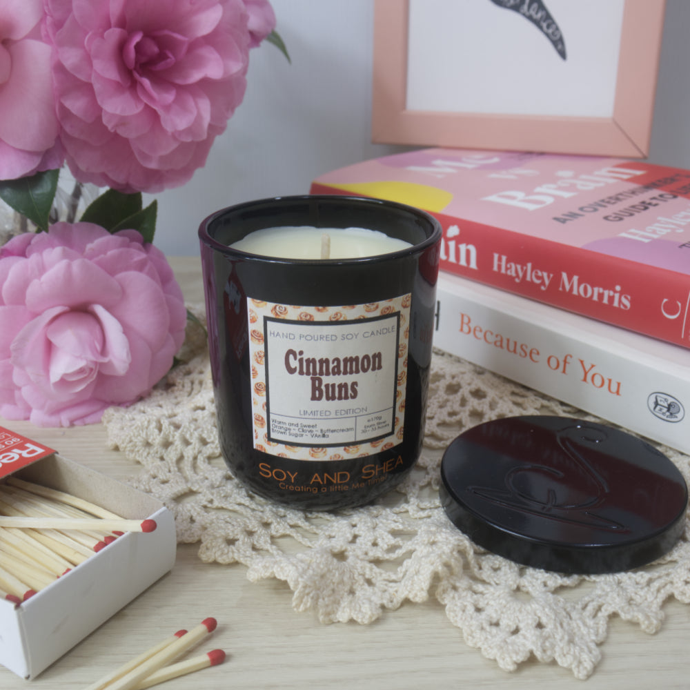 A black candle jar with decorative label is on a sideboard with vase of pink flowers to the left and a stack of bookes to the right.   The lid of the jar has been removed and placed to the right in front of the books revealing a cream wax.