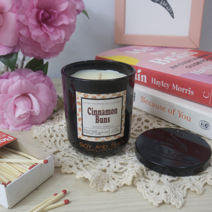 A black candle jar with decorative label is on a sideboard with vase of pink flowers to the left and a stack of bookes to the right.   The lid of the jar has been removed and placed to the right in front of the books revealing a cream wax.